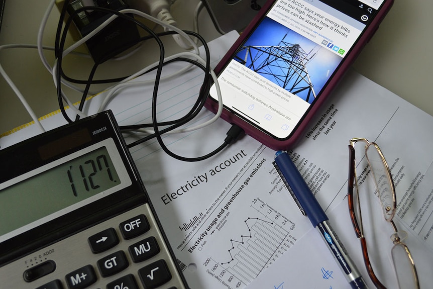 A flat lay of a calculator, an electricity bill and a mobile phone displaying an ABC article about electricity prices. 