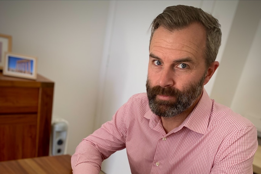 A man in a pink shirts sits on a table with a laptop.