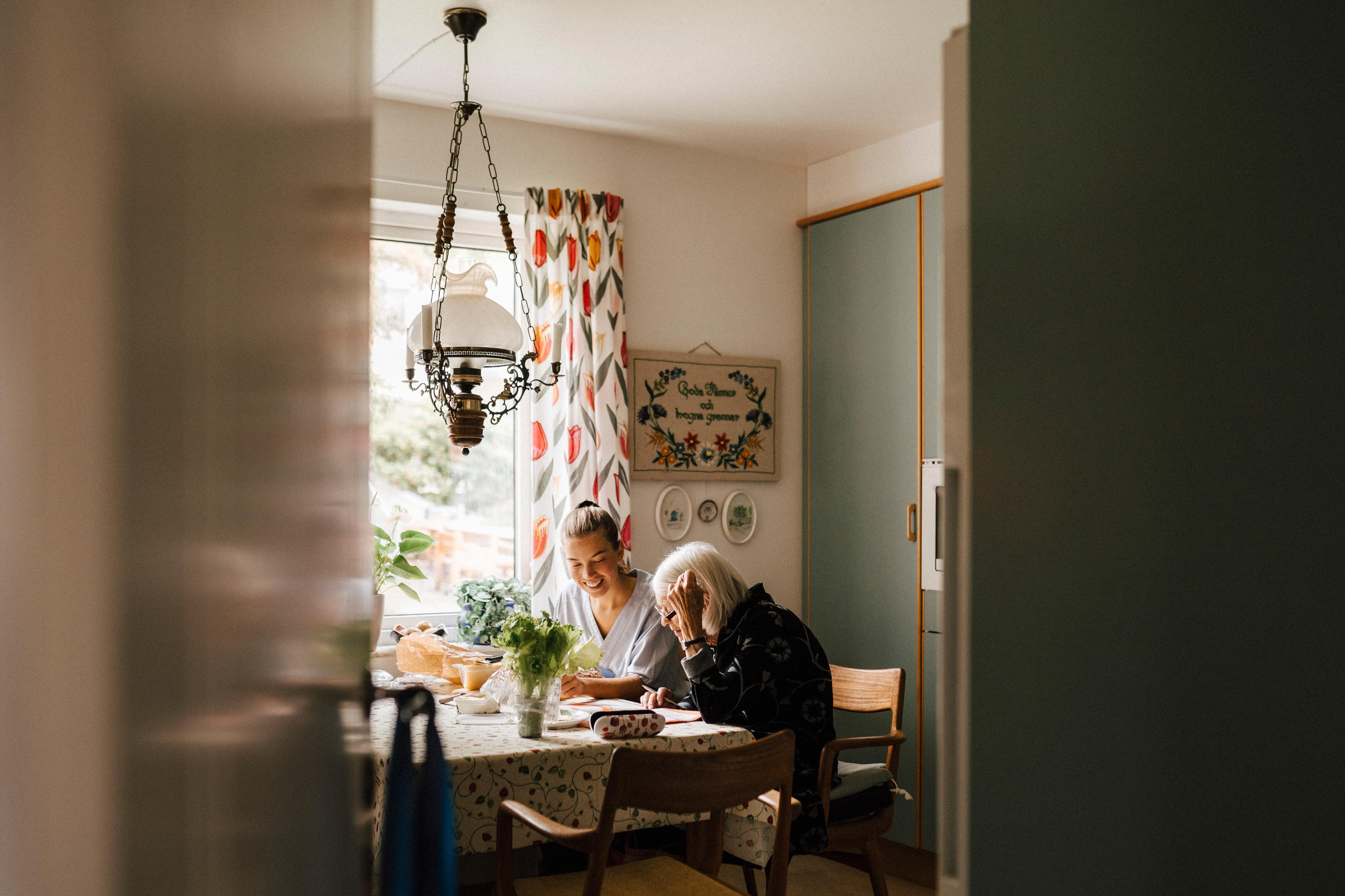 Senior woman reading paper sitting by female caregiver in kitchen at home
