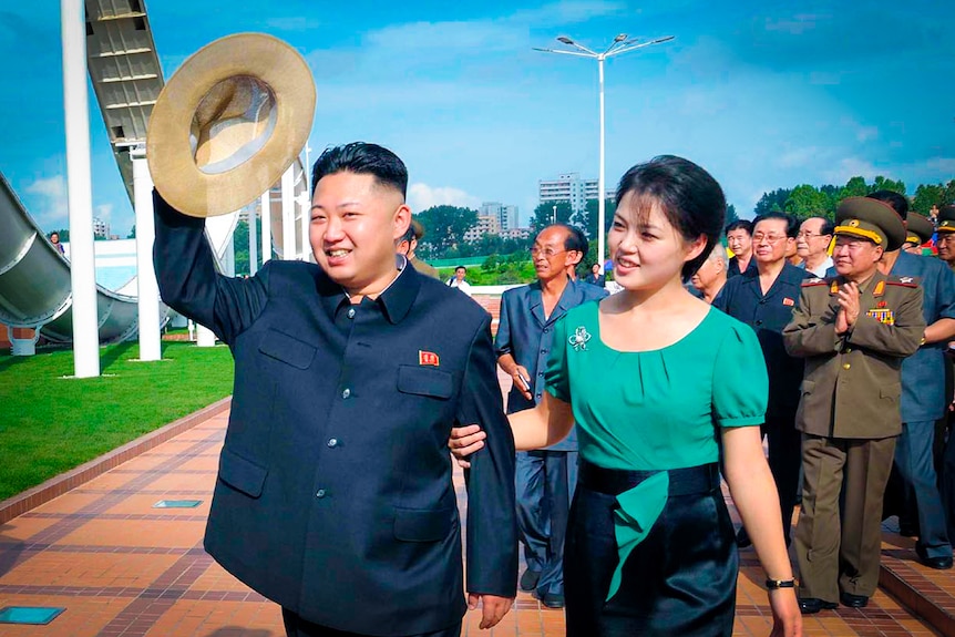 Kim Jong Un waves around a big straw hat, while a woman in a green dress holds his arm and smiles