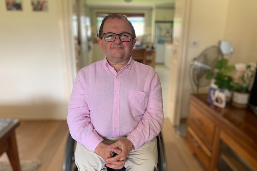 A middle-aged man sitting in a wheelchair. He's wearing glasses and a pink shirt. 