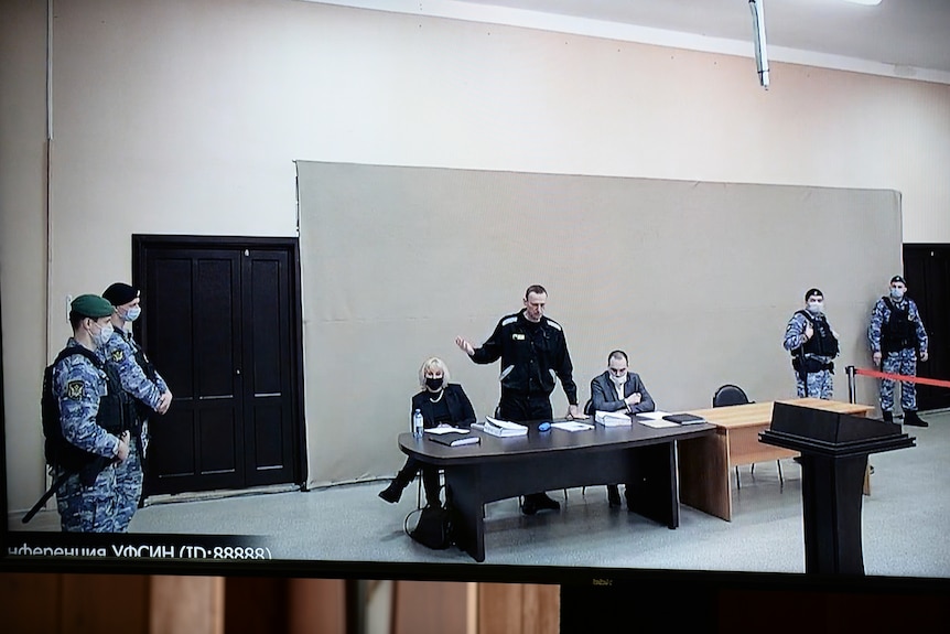 Mężczyzna rozmawia przez wideo, stojąc obok swojego adwokata w osobnej sali sądowej.