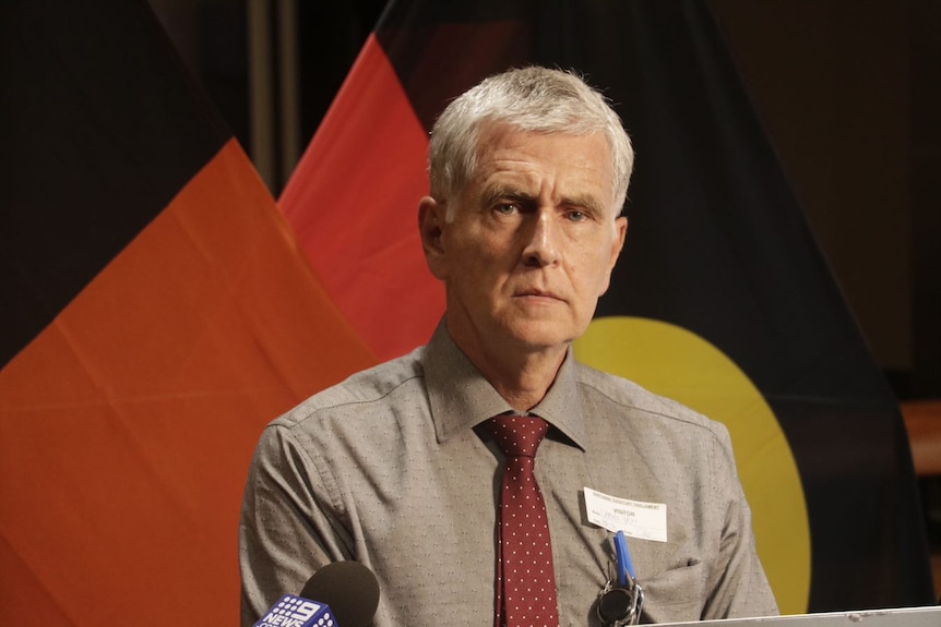 A man looks sternly at the camera in front of an Aboriginal flag. 