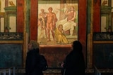 Two people stand underneath a lit Roman mural hanging on a wall. 