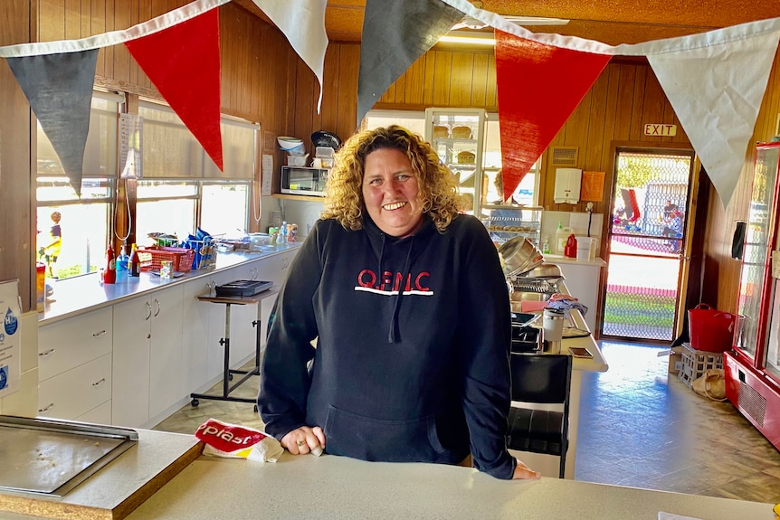 A woman with curly red hair stands behind the counter of a canteen.  There are red, white and black streamers above her head.