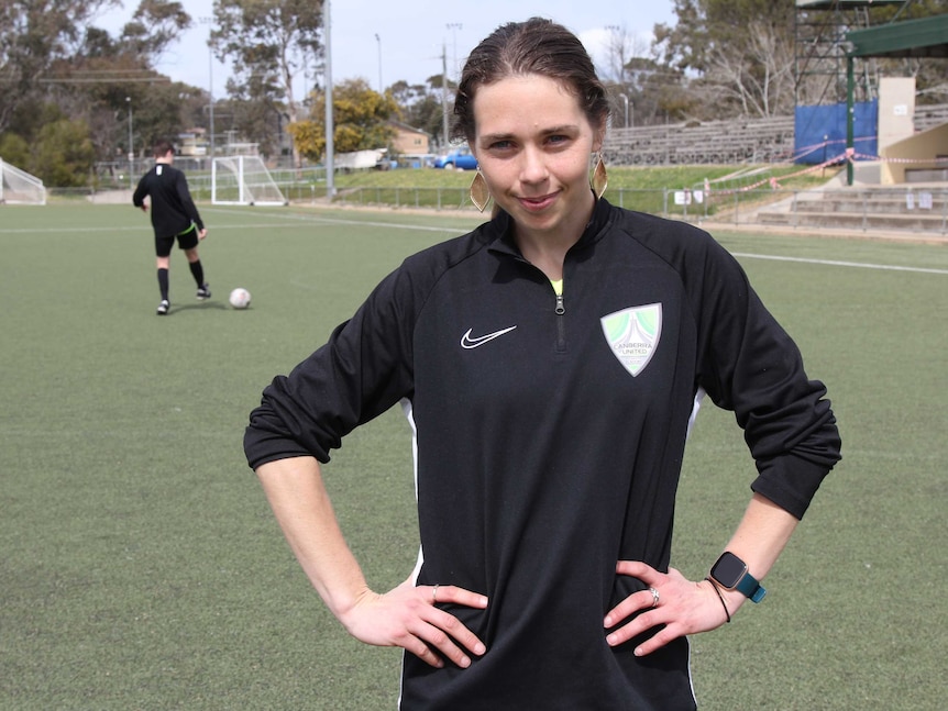 A teenage girl stands smiling, in a soccer jersey on a football pitch.