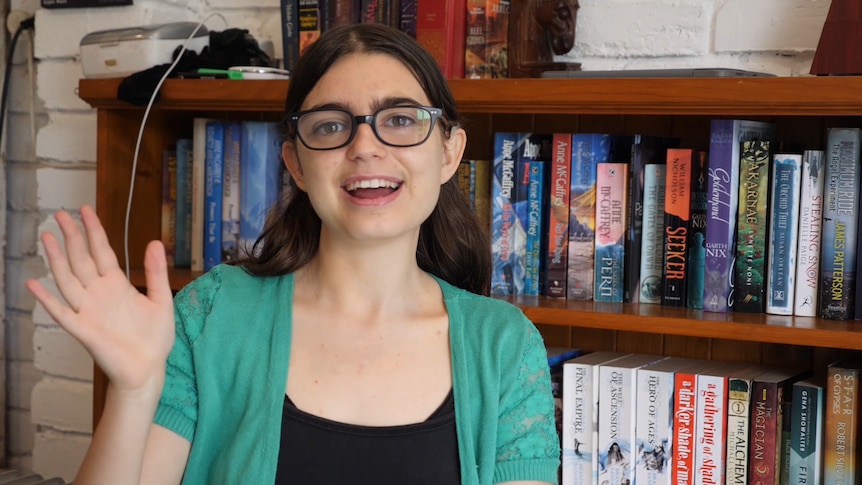 Tallula Bourne waves at the camera. She wears a green top & glasses and sits in front of a bookshelf filled with fantasy novels.