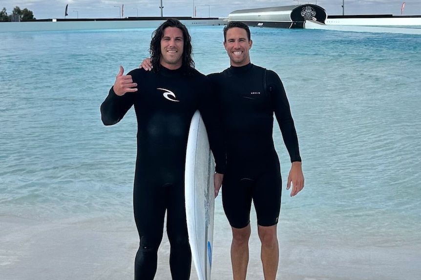 Two men in wetsuits stand together near the water