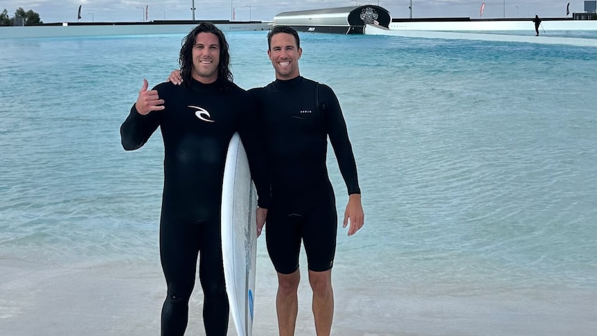 Two men in wetsuits stand together near water