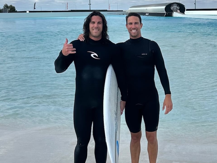 Two men in wetsuits stand together near water