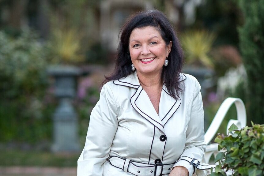 Dark-haired woman in white trench coat walks through a garden smiling at the camera.