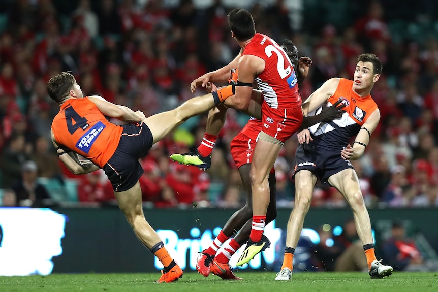 A GWS AFL player stands on one leg while holding the ball and putting his foot out to block a Swans player trying to tackle him.