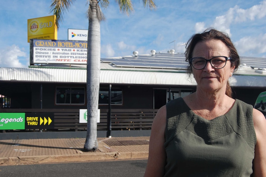 A woman stands out the front of a pub with the sign 'Grand Hotel-Motel'. A palm tree is behind her.