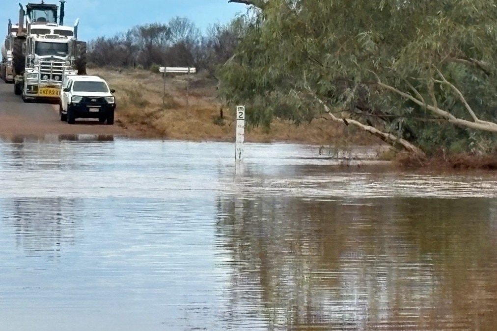 Uma estrada alagada, com um ute e um caminhão parados ao fundo. Um estadiômetro mede água a uma altura de 1 metro.