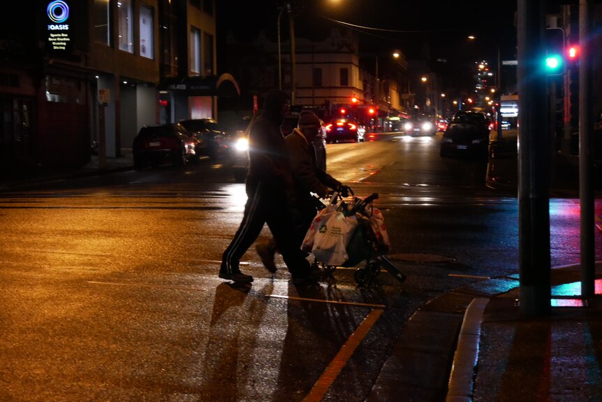 Two people cross a street in the dark pushing a walker covered in plastic shopping bags.