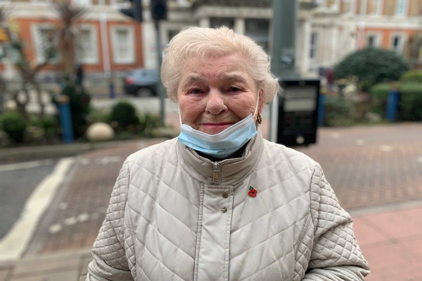 An elderly woman stands in a street a facemask hanging below her mouth.