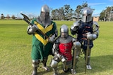 Three men wear full knight's armour. Left has green and yellow coat holding axe, centre red and right blue holding swords.