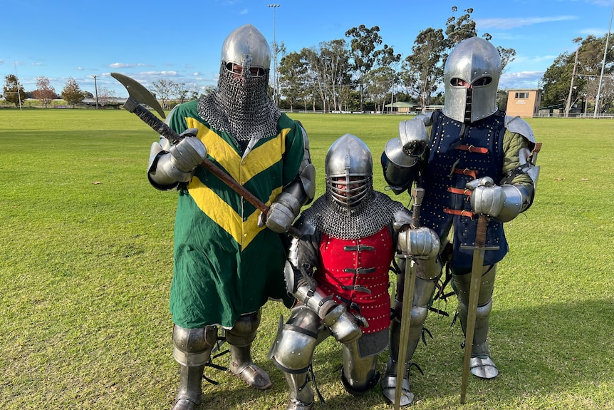 Three men wear full knight's armour. Left has green & yellow coat holding axe, centre red & right blue holding swords.