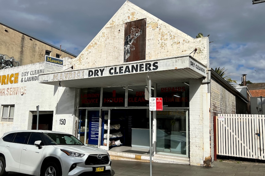 A photo from the street of an aging white brick single story building with 'Maurice Drycleaners' printed on it in black letters.