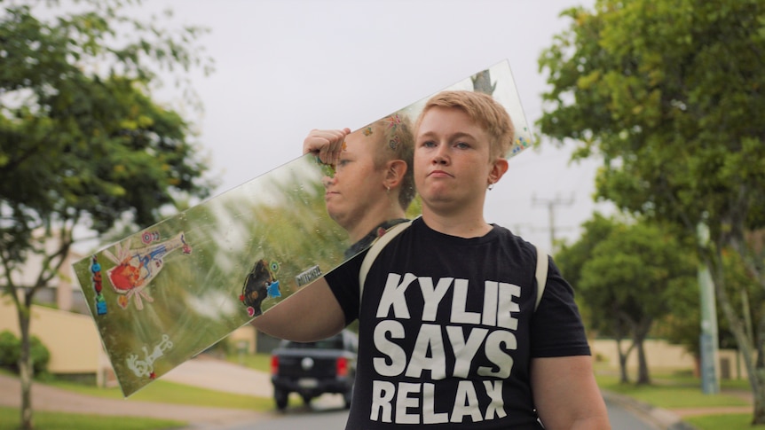Kylie walks down the street holding a long mirror over her shoulder. She is wearing a tshirt saying Kylie Says Relax