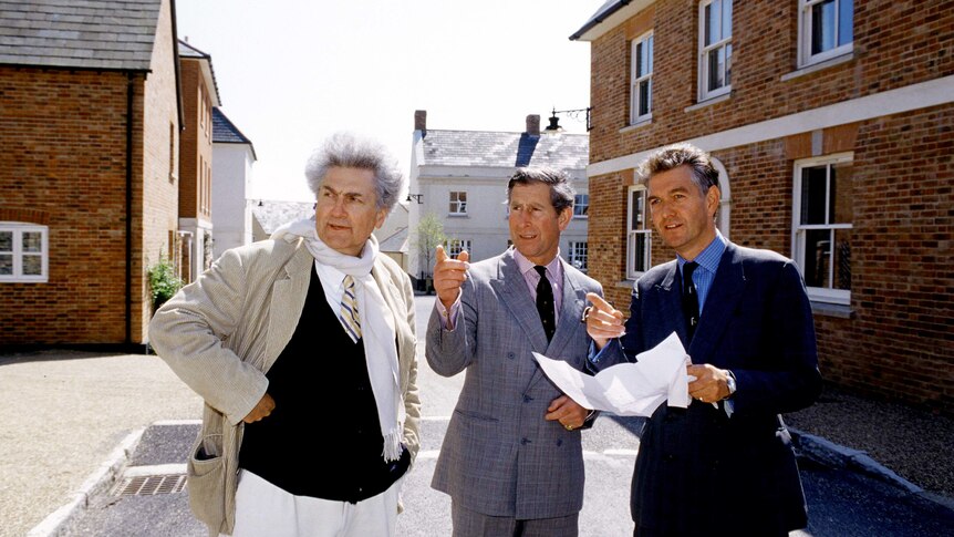 On a bright day on a neo-Georgian street, you view Prince Charles pointing into the distance with two men beside him.
