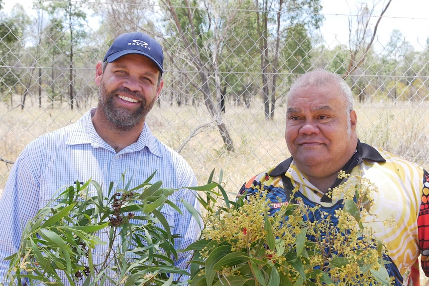 Two man standing. One is wearing a cap and cheque shirt smiling. The other is wearing a polo shirt. Both are holding wattle