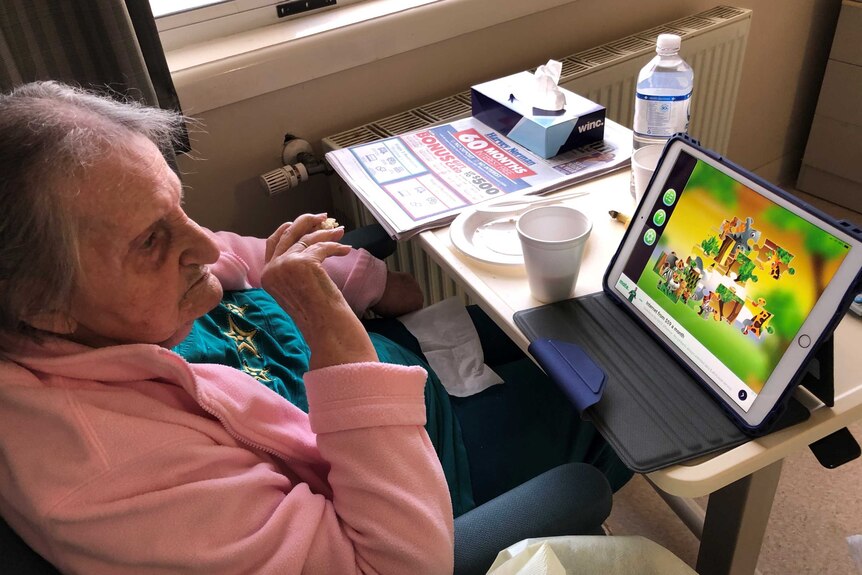 An elderly woman watches a puzzle game on an Ipad screen as she eats a meal.