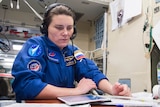 A woman, Anna Kikina, is wearing a blue overall and headphones with a microphone, while looking at papers in front of her.