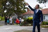 An auctioneer raises his arm, standing on a suburban street in front of a small crowd of people.