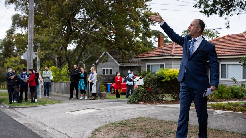 An auctioneer raises his arm, standing on a suburban street in front of a small crowd of people.