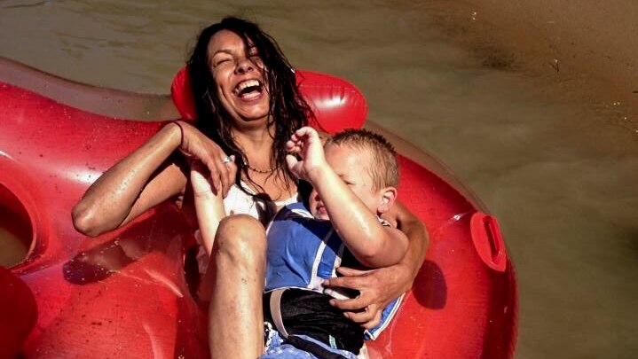 Tanya Day, laughing, laying on an inflatable device on a brown river, holding a small child.