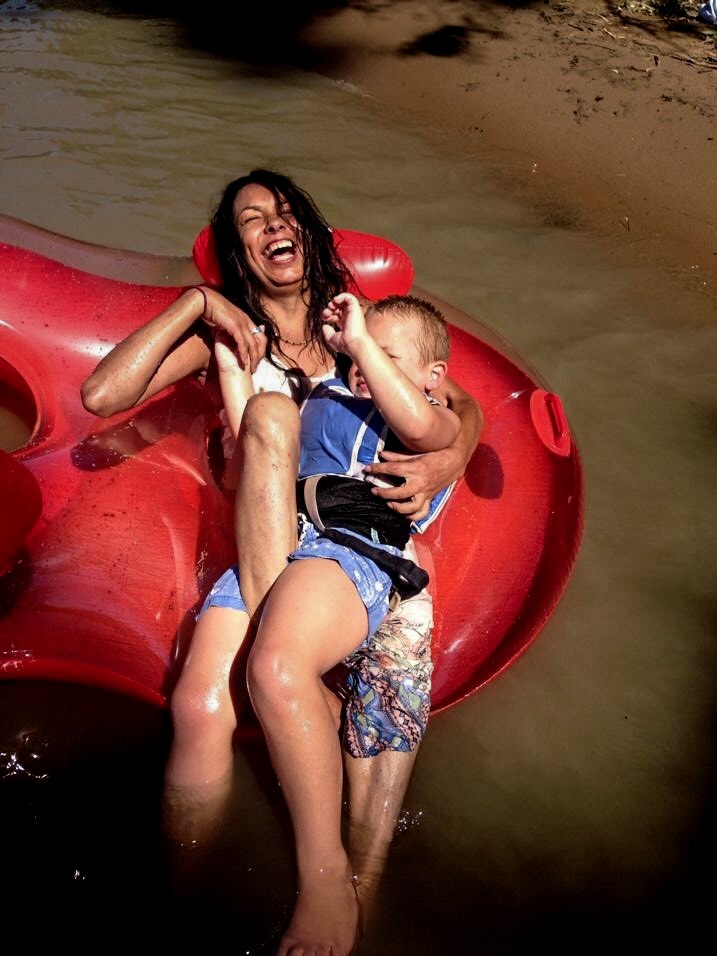 Tanya Day, laughing, laying on an inflatable device on a brown river, holding a small child.