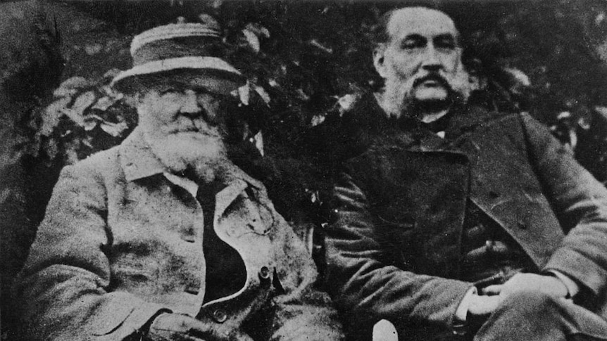 Old black and white photograph of a middle aged formally dressed man with a large mustache in a garden next to a older man 