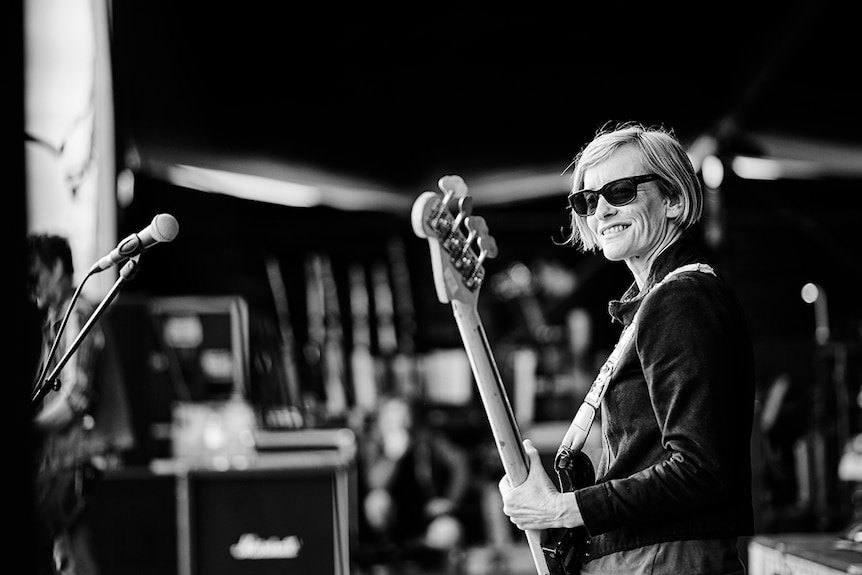 Black and white photo of Janet English playing bass guitar on stage. She wears sunglasses and a jacket.