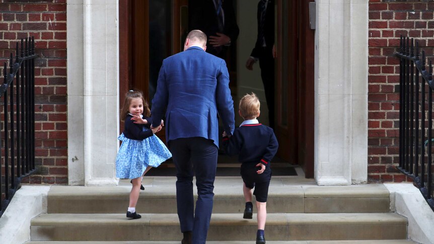 Prince William, Prince George and Princess Charlotte walk upstairs into a hospital with Princess Charlotte waving.
