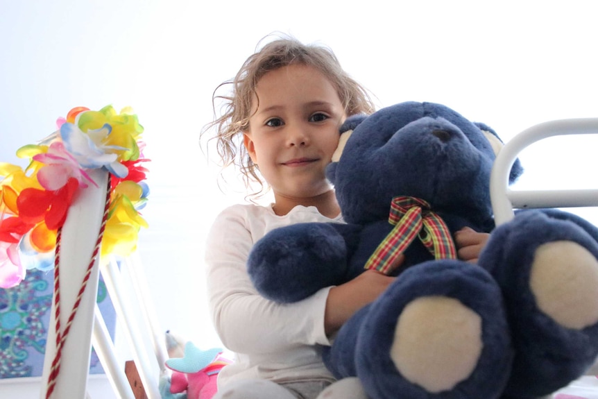 Zara cuddles her teddy and looks into the camera, there are bright flowers on the other side of her.