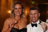 A woman in a black dress and a man in a white suit and black tie hold up medals and smile