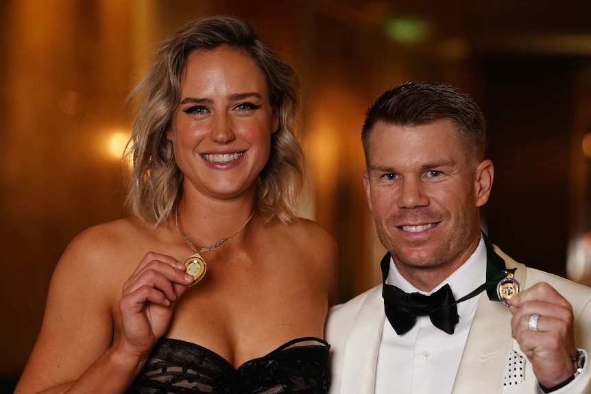 A woman in a black dress and a man in a white suit and black tie hold up medals and smile