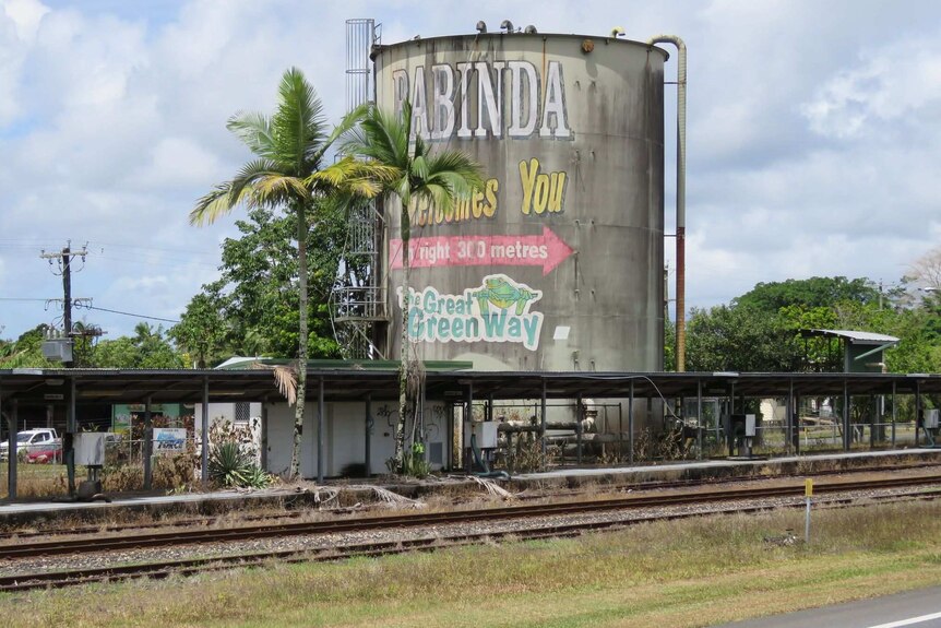 Rusting and discoloured molasses tank with ladder and pipes coming from it with Babinda Welcomes You in faded paint on the side