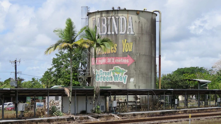 Rusting and discoloured molasses tank with ladder and pipes coming from it with Babinda Welcomes You in faded paint on the side