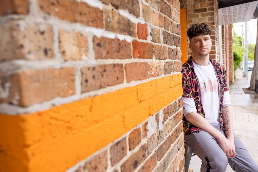 White man with curly cropped brown hair wears red and black chain shirt over white t-shirt and leans against brick wall.