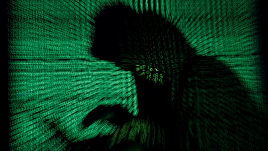 A hooded man holds a laptop computer as cyber code is projected on him 