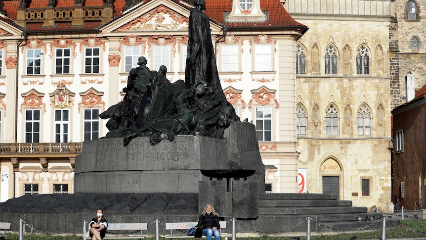 Two women with masks sit on a bench keeping social distance at the Old Town Square