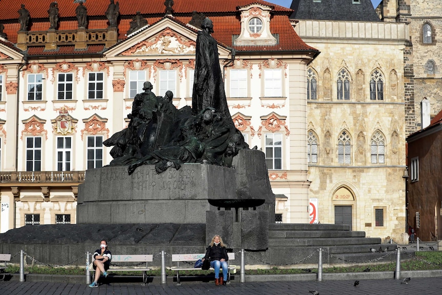 Two women with masks sit on a bench keeping social distance at the Old Town Square