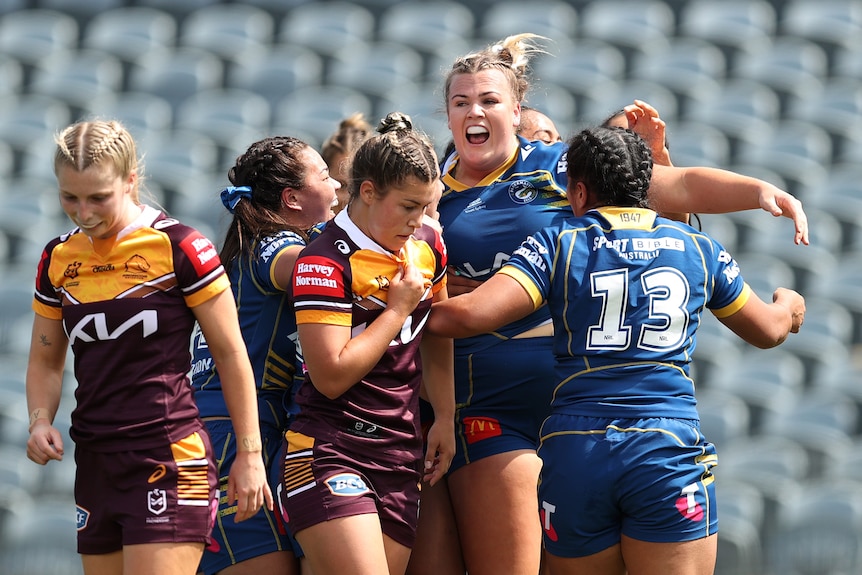 An Eels NRLW player stands calling out in celebration as her teammates come in past dejected Broncos players.