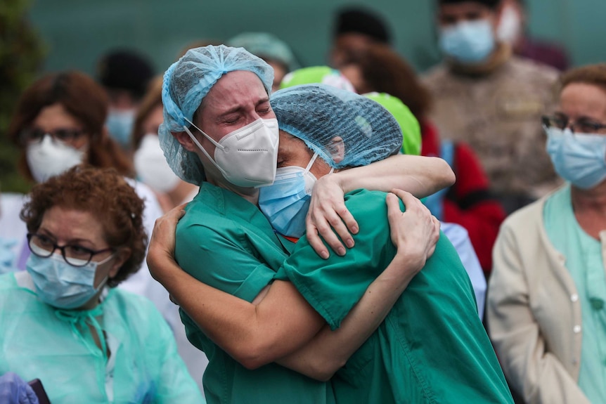 Two women in hairnets, masks and green nursing scrubs embrace and cry.