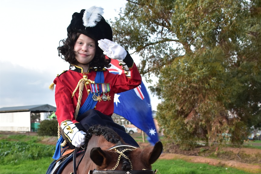 A young girl sits on a horse dressed up in formal military costume and salutes