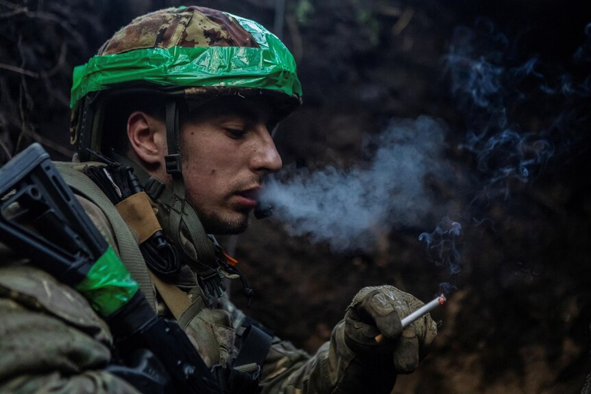 A solider holds a cigarette as he sits in a dark trench. 
