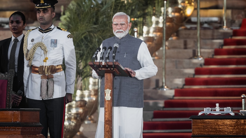 Narendra Modi standing at a lectern with mics and staircase with red carpet in the background
