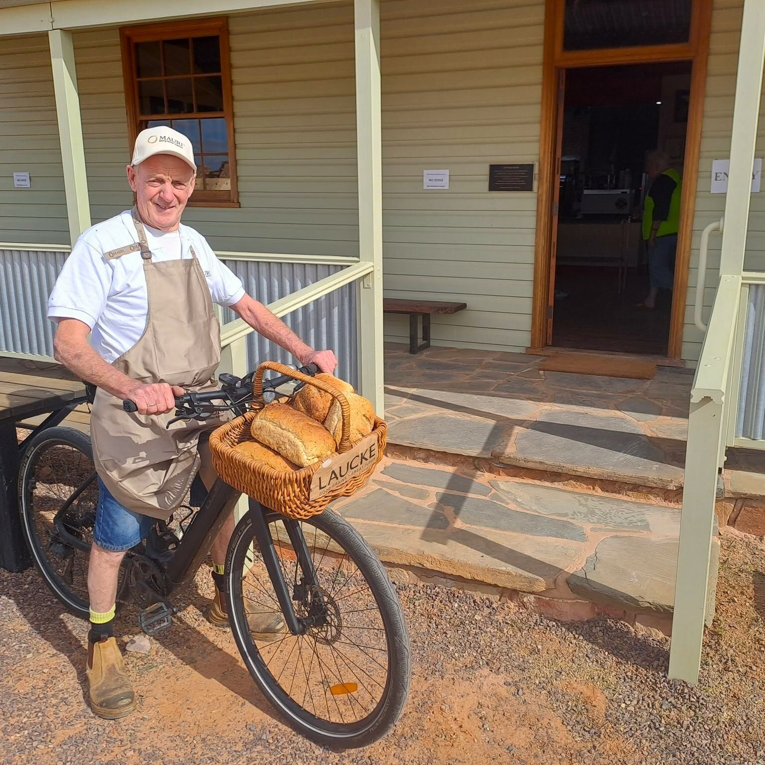 A man on a bike with bread in a basket outside a weatherboard cafe.